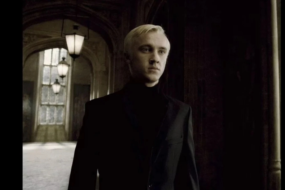 Том Фелтон, актер, сыгравший Драко Малфоя, улыбается, на фоне стилизованного изображения Хогвартса и символов Гарри Поттера, указывая на постер фильма «Гарри Поттер и Тайная комната»